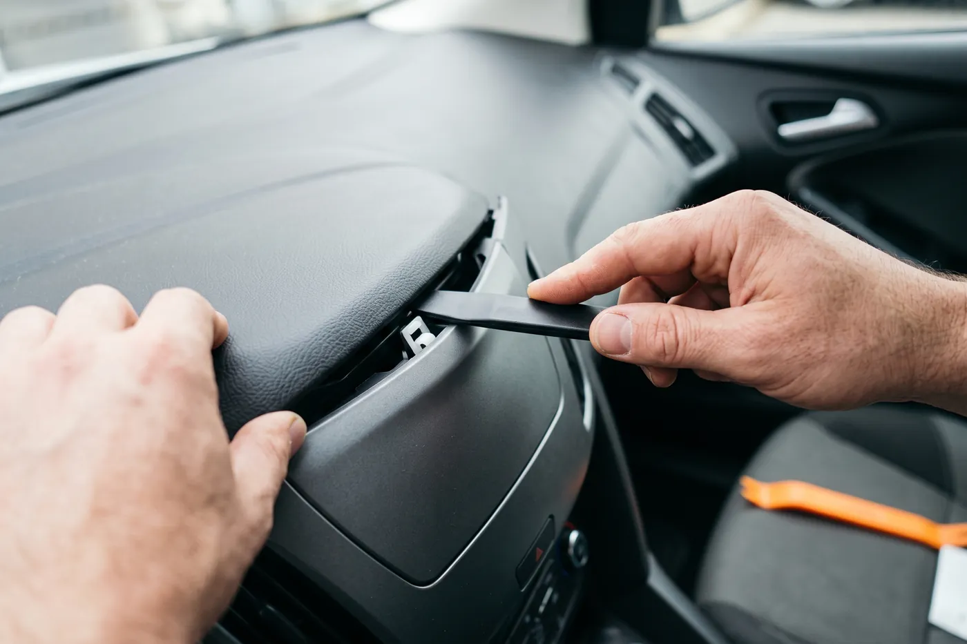 Hands using a plastic trim tool to lever a dashboard bezel away from the centre console, with the clips beginning to release.